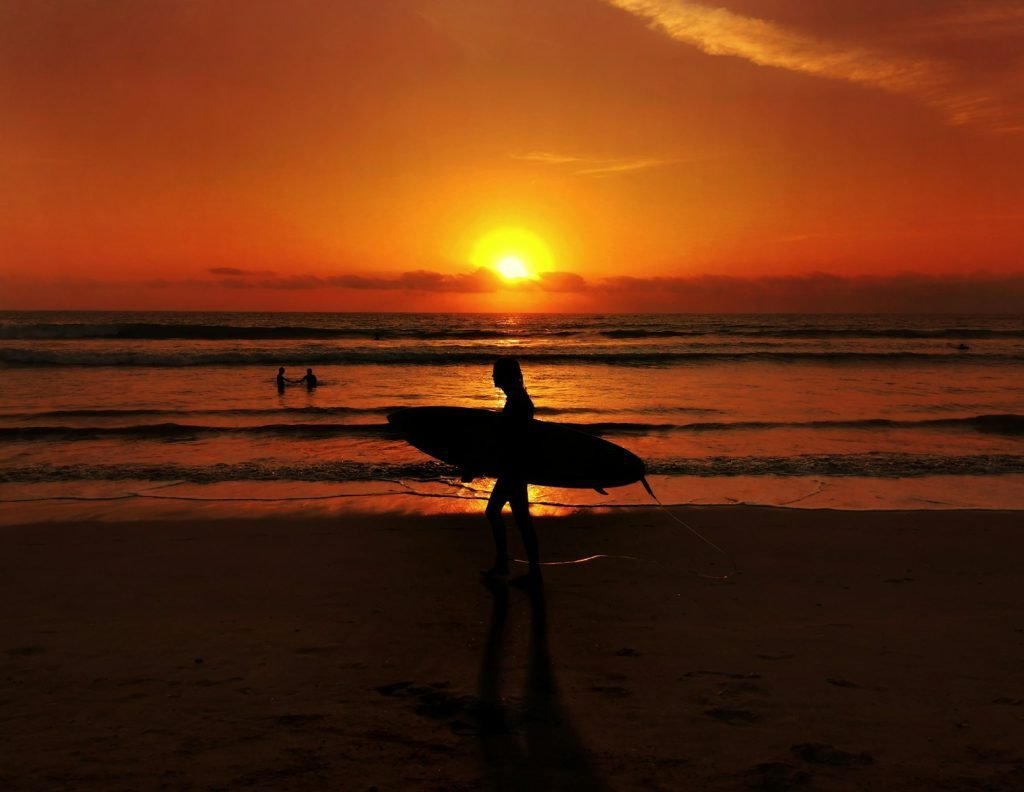 silhouette of man holding surfboard walking on beach during sunset kuta