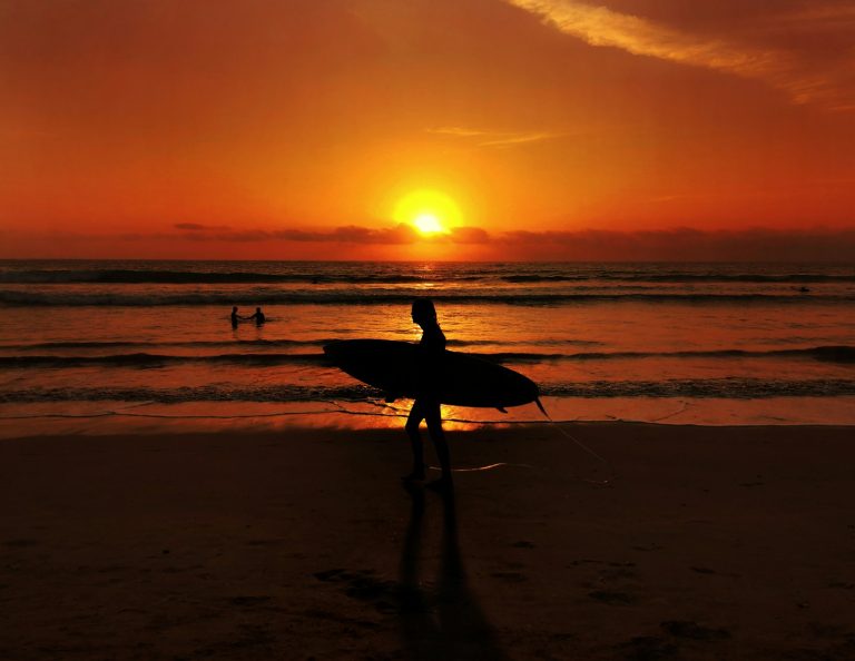 silhouette of man holding surfboard walking on beach during sunset kuta