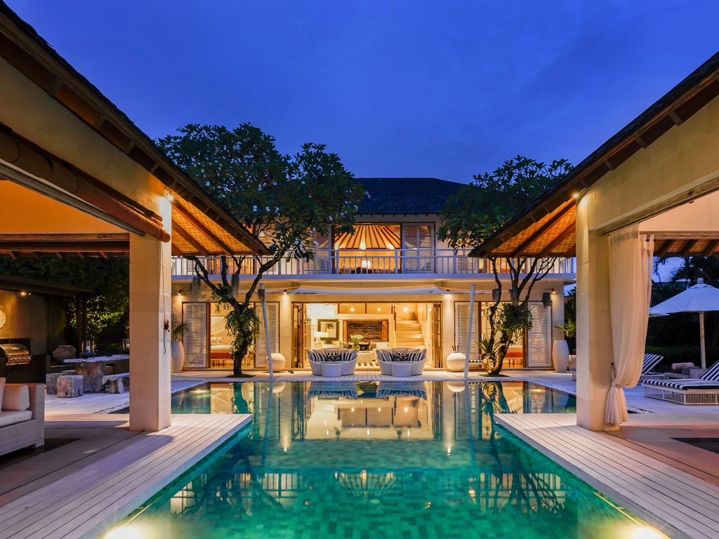Villa Jajaliluna Seminyak courtyard pool villa at dusk with glowing lights open living spaces and trees around the water