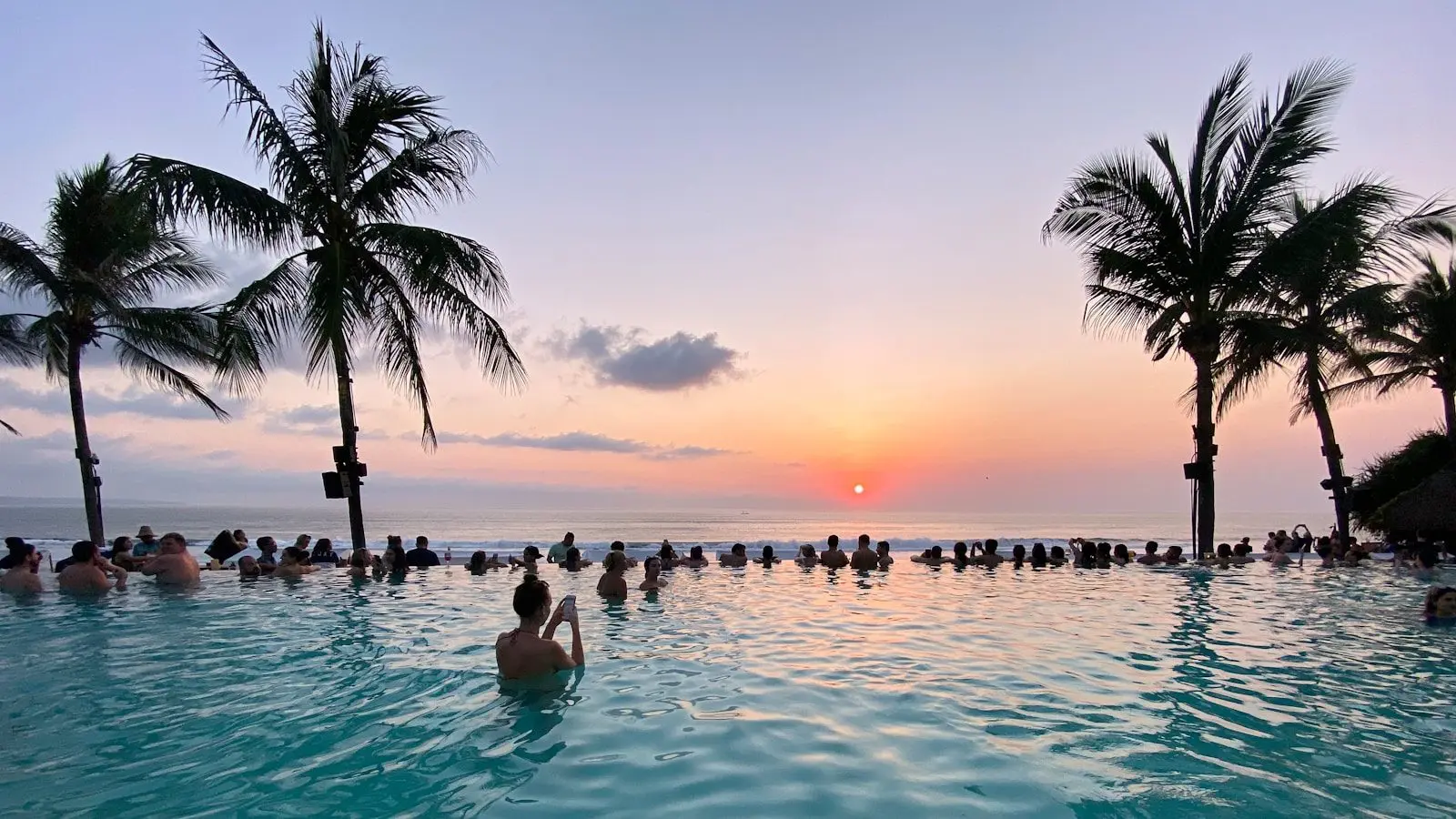 Kolam renang dan pohon kelapa menghadap laut saat matahari terbenam di Potato Head Beach Club Seminyak